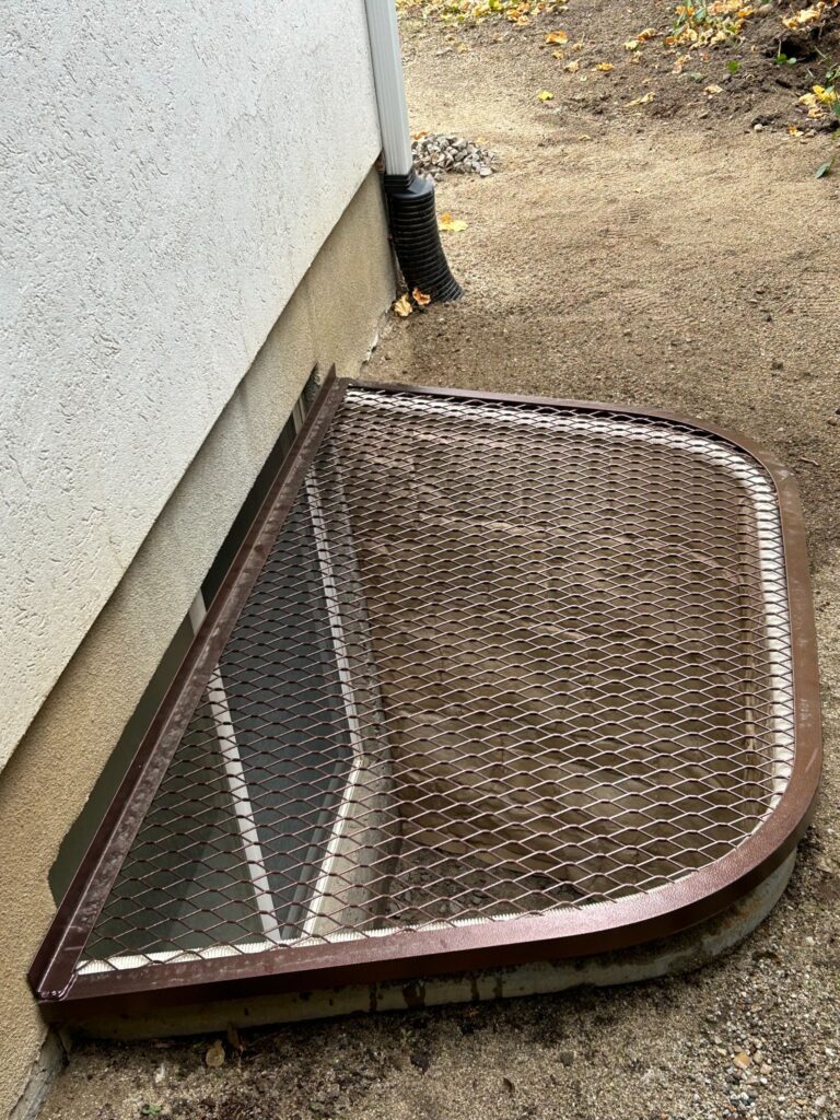 A brown steel mesh window well grate fitted over a basement egress window well next to a house foundation.