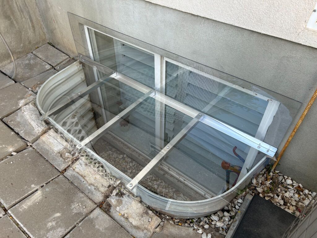 A clear polycarbonate basement window well cover supported by metal support bars installed over a metal window well.
