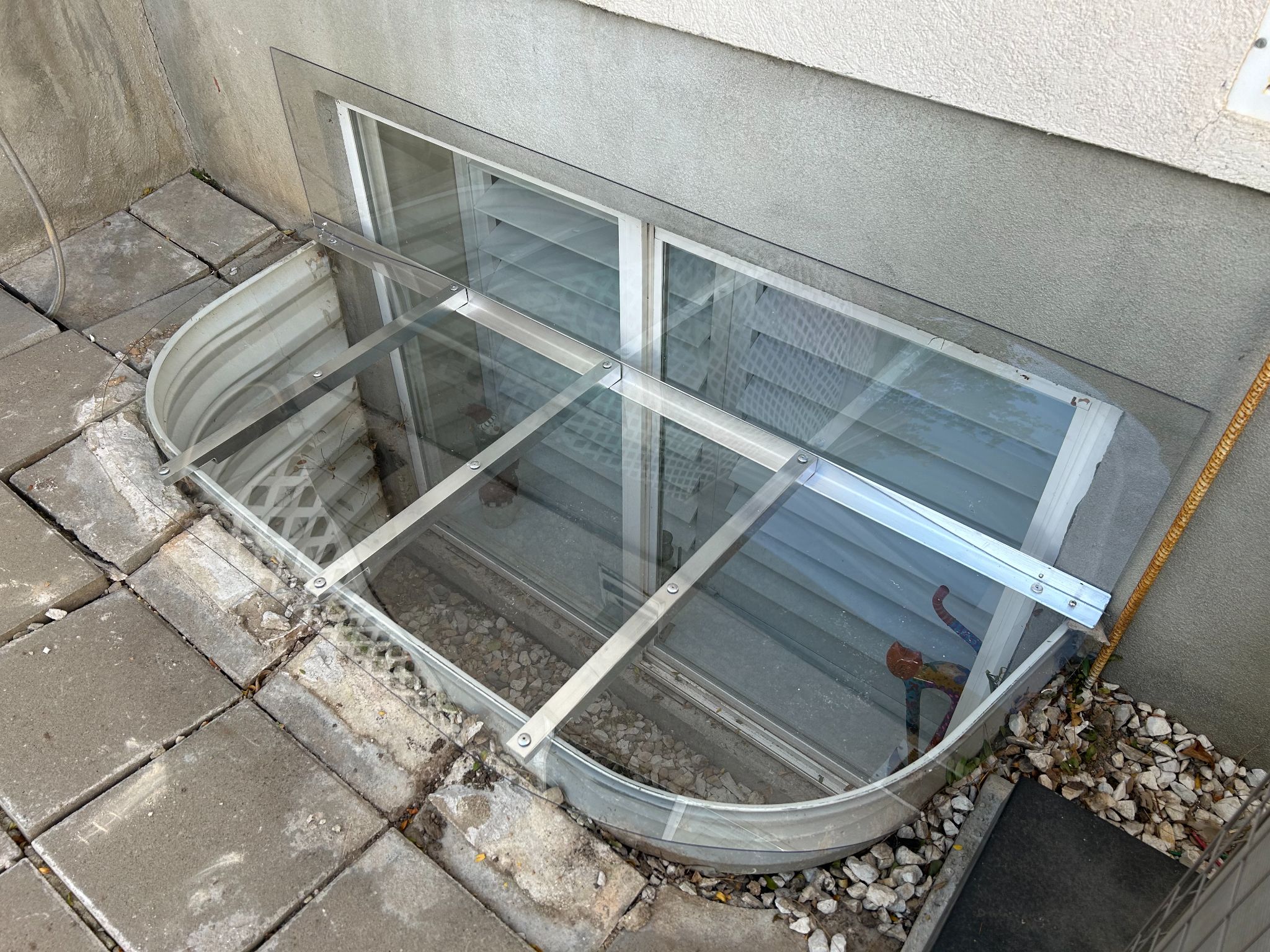 A clear polycarbonate basement window well cover supported by metal support bars installed over a metal window well.
