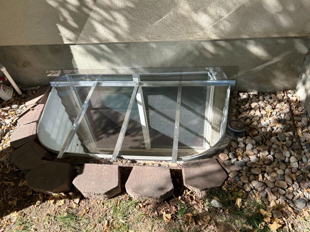 Clear window well cover with metal support bars installed over a basement egress window surrounded by stone pavers.