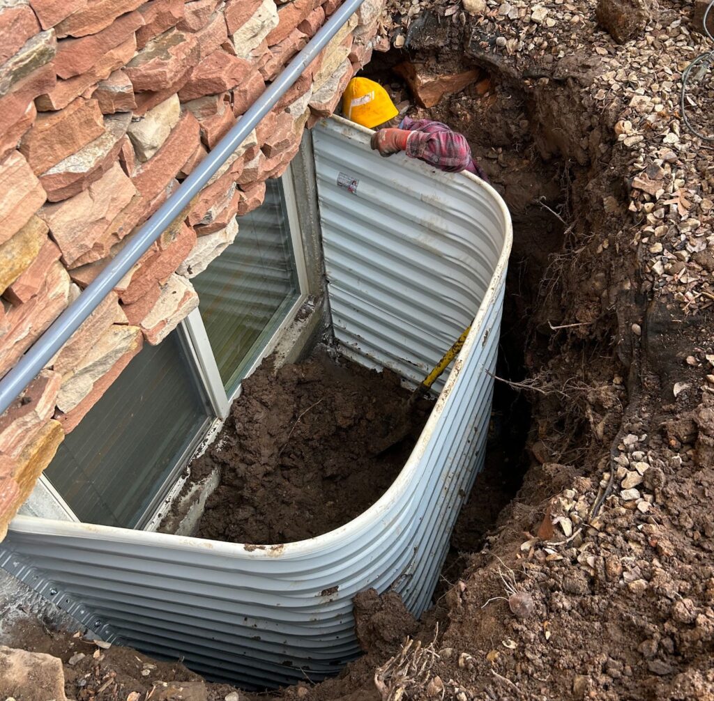 A construction worker in a yellow hard hat installs a corrugated metal window well against a brick exterior wall, surrounded by excavated soil and roots.