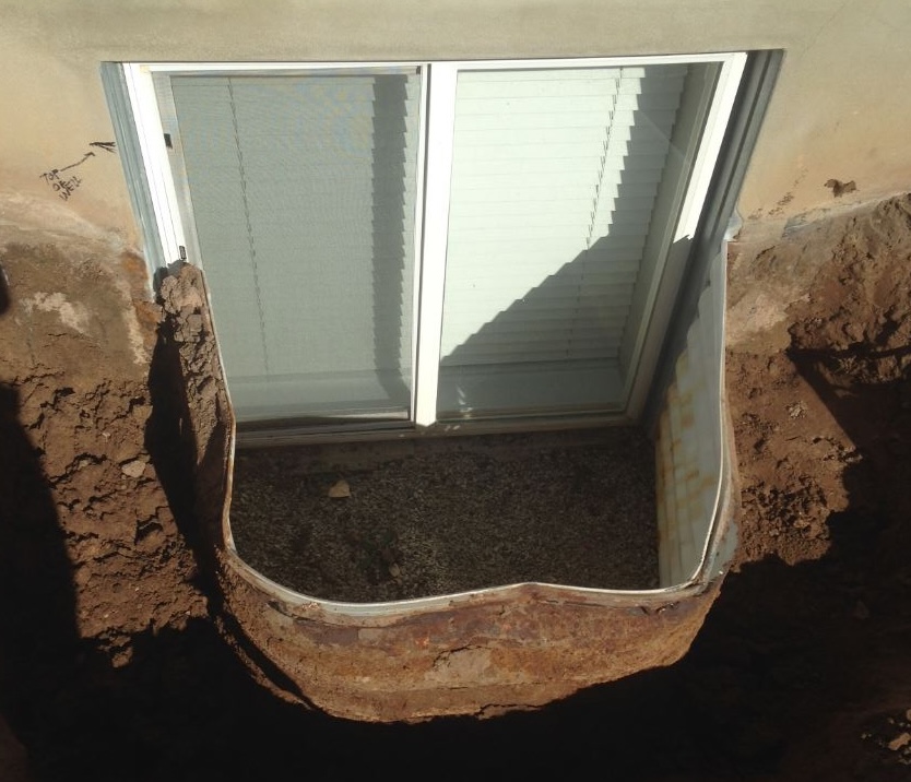 Basement window with blinds in a rusty corrugated metal well filled with gravel, surrounded by excavated dirt.