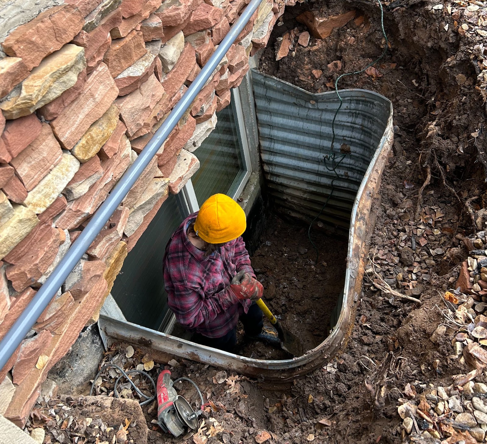 Person in a yellow hard hat and plaid jacket digging in a metal-framed window well next to a brick wall.