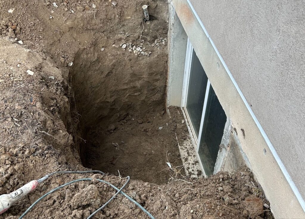 The image shows an excavated area next to a building foundation, with a window well exposed and some cables or pipes visible in the dirt.