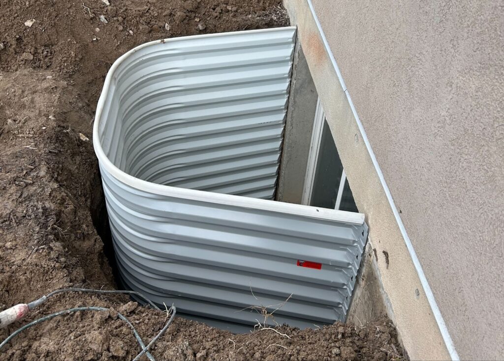 This image shows a basement window well installation, featuring a corrugated metal window well attached to the exterior wall of a building, with surrounding excavated soil.
