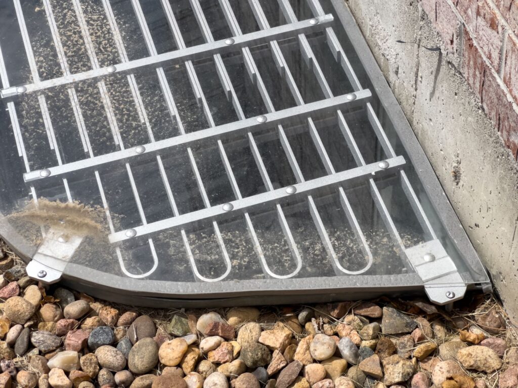 A metal grate on the ground beside a brick wall, designed as a custom window well cover for safety and ventilation.