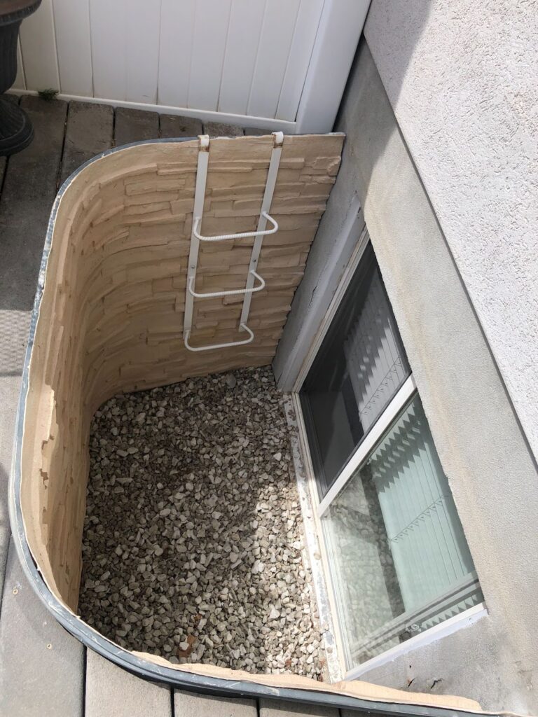 Basement window well with gravel floor, stone-look liner, white metal escape ladder hanging on wall, and egress window partially open. Outdoor access point.
