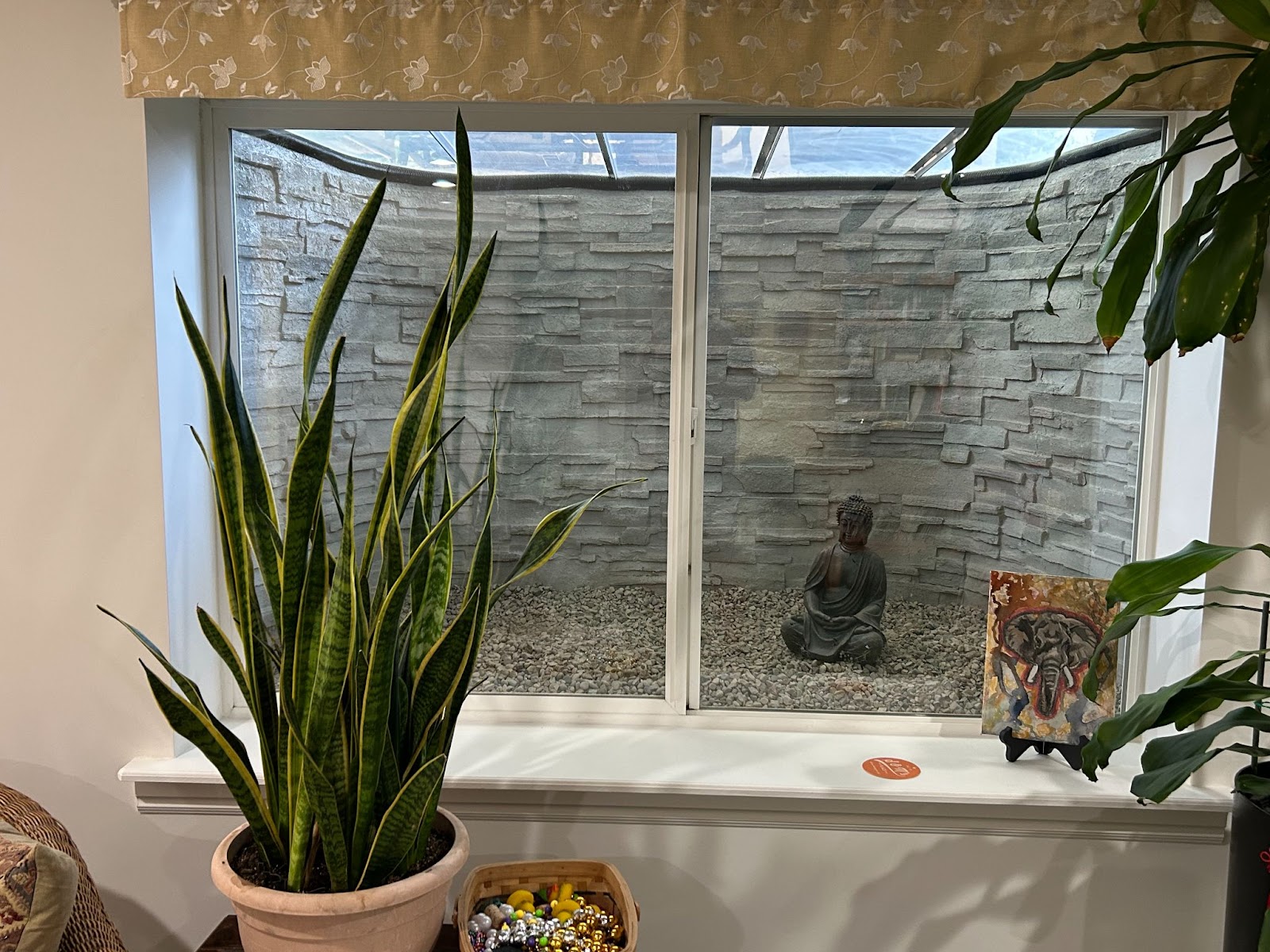 Zen garden window with Buddha statue, pebbles, snake plant, elephant art, and peace lily. Cozy indoor meditation nook.