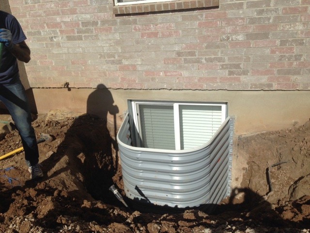 A worker installing a corrugated metal window well around a basement window in an excavated area.