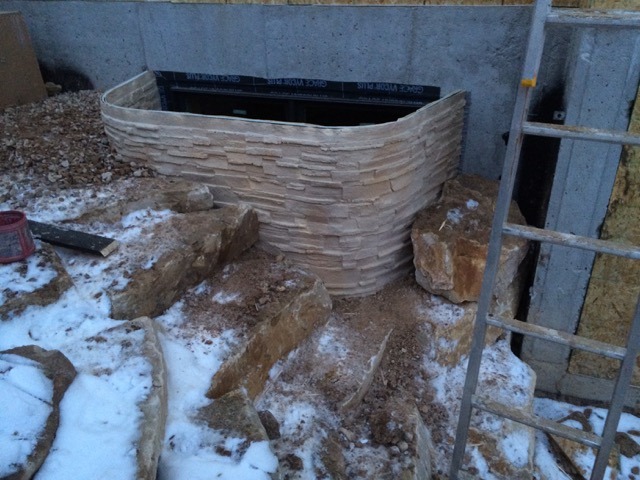 A decorative, stone-textured window well installed around a basement window in a snowy, excavated area.