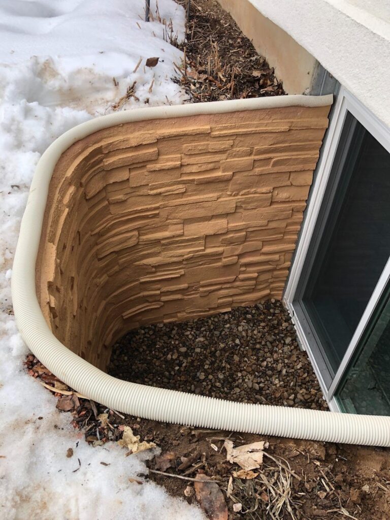 A small window on the side of a house featuring a drain below it for water runoff