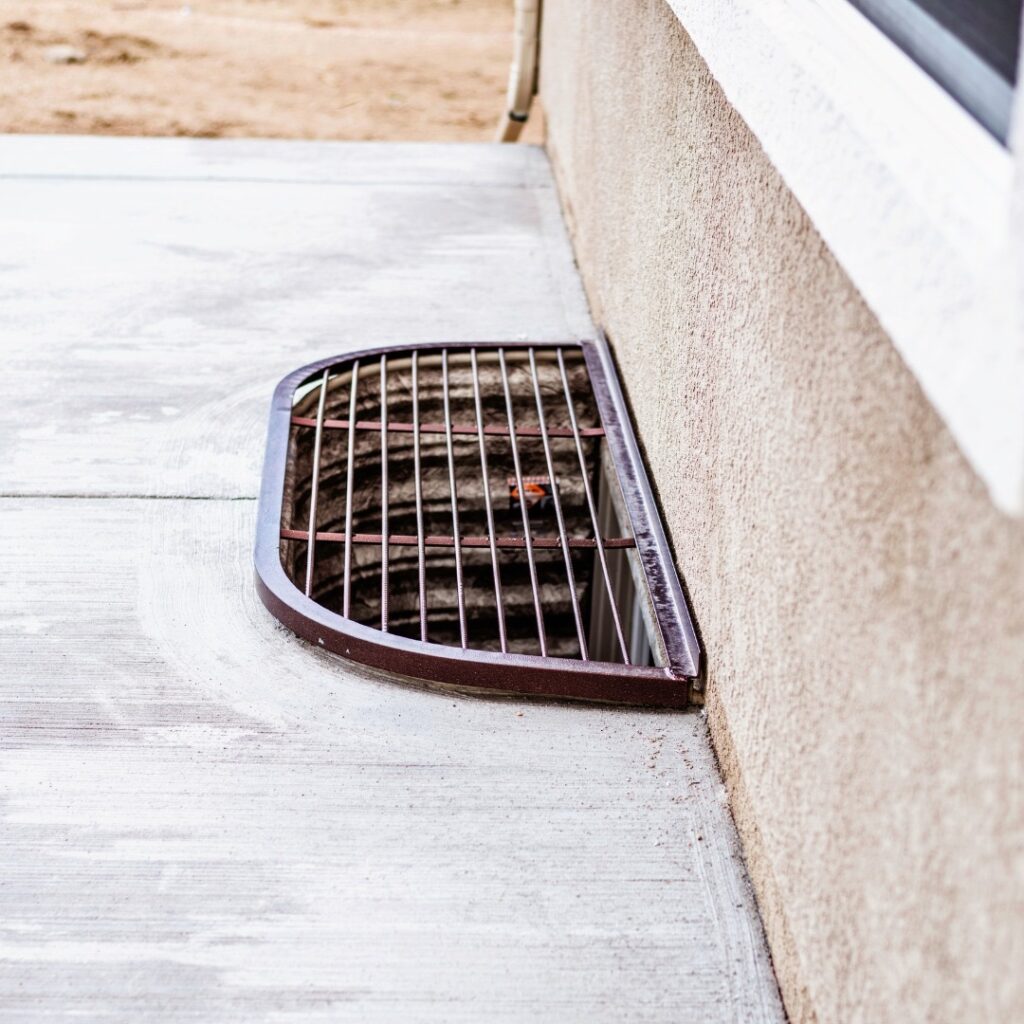 A metal grate cover placed over a semi-circular window well flush with a concrete patio.
