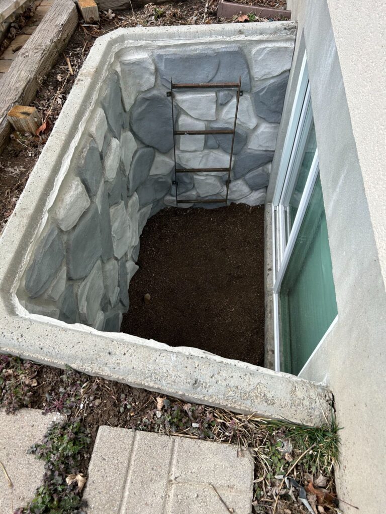 Concrete egress window well with a decorative grey stone-patterned liner and a built-in metal escape ladder.