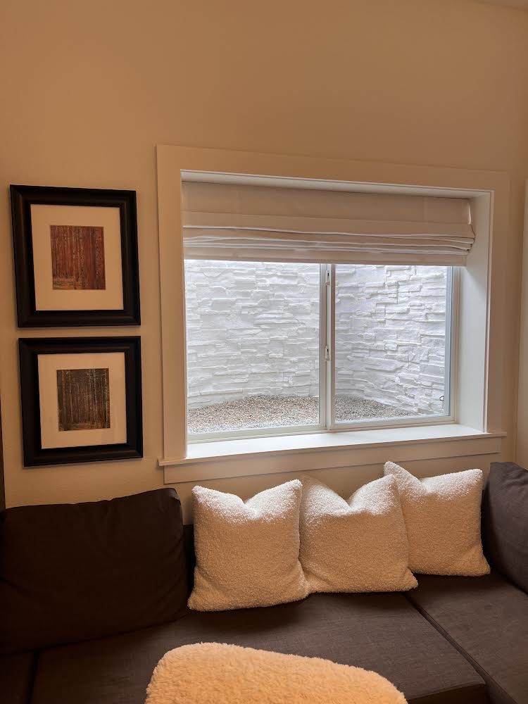 A basement window looking out into a white stone window well, with three white textured throw pillows lined up on a brown sectional sofa below it and two framed art pieces on the left.