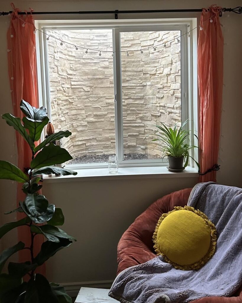 A cozy room corner featuring a basement window with a stone window well, framed by pink curtains and string lights, with a round chair holding a yellow pillow and grey blanket in the foreground.