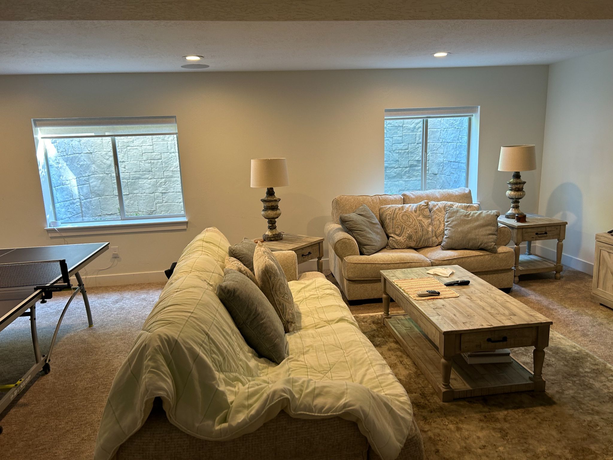 A furnished basement living room featuring two beige sofas with throw pillows, a wooden coffee table, two side lamps, and a ping pong table on the left near a window.